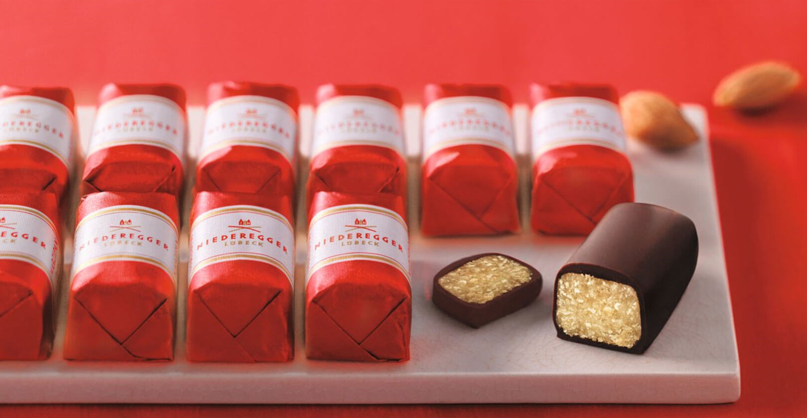 Marzipan Klassiker der Firma Niederegger in zwei Reihen aufgereiht, eingepackt in rote Wickler mit einer weißen Banderole. Auf der rechten Seite ist ein Klassiker angeschnitten worden.