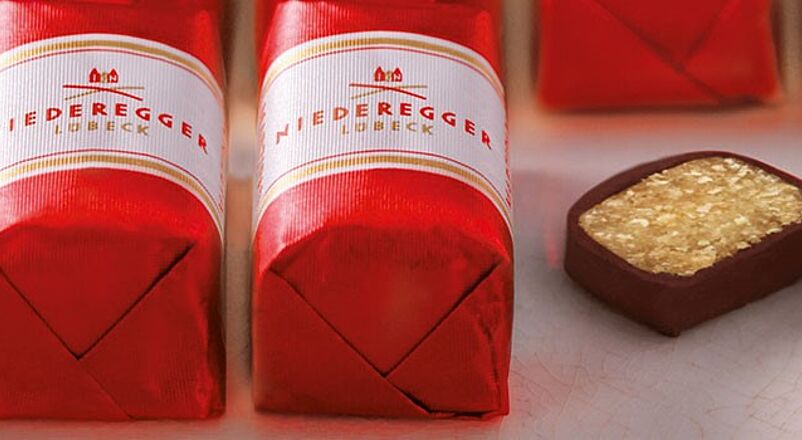 Marzipan Klassiker der Firma Niederegger in zwei Reihen aufgereiht, eingepackt in rote Wickler mit einer weißen Banderole. Auf der rechten Seite ist ein Klassiker angeschnitten worden.