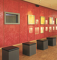 Innenansicht des Niederegger Marzipanmuseums. An der roten Wand hängen verschiedene Ausstellungsstücke mit Sitzgelegenheiten davor.